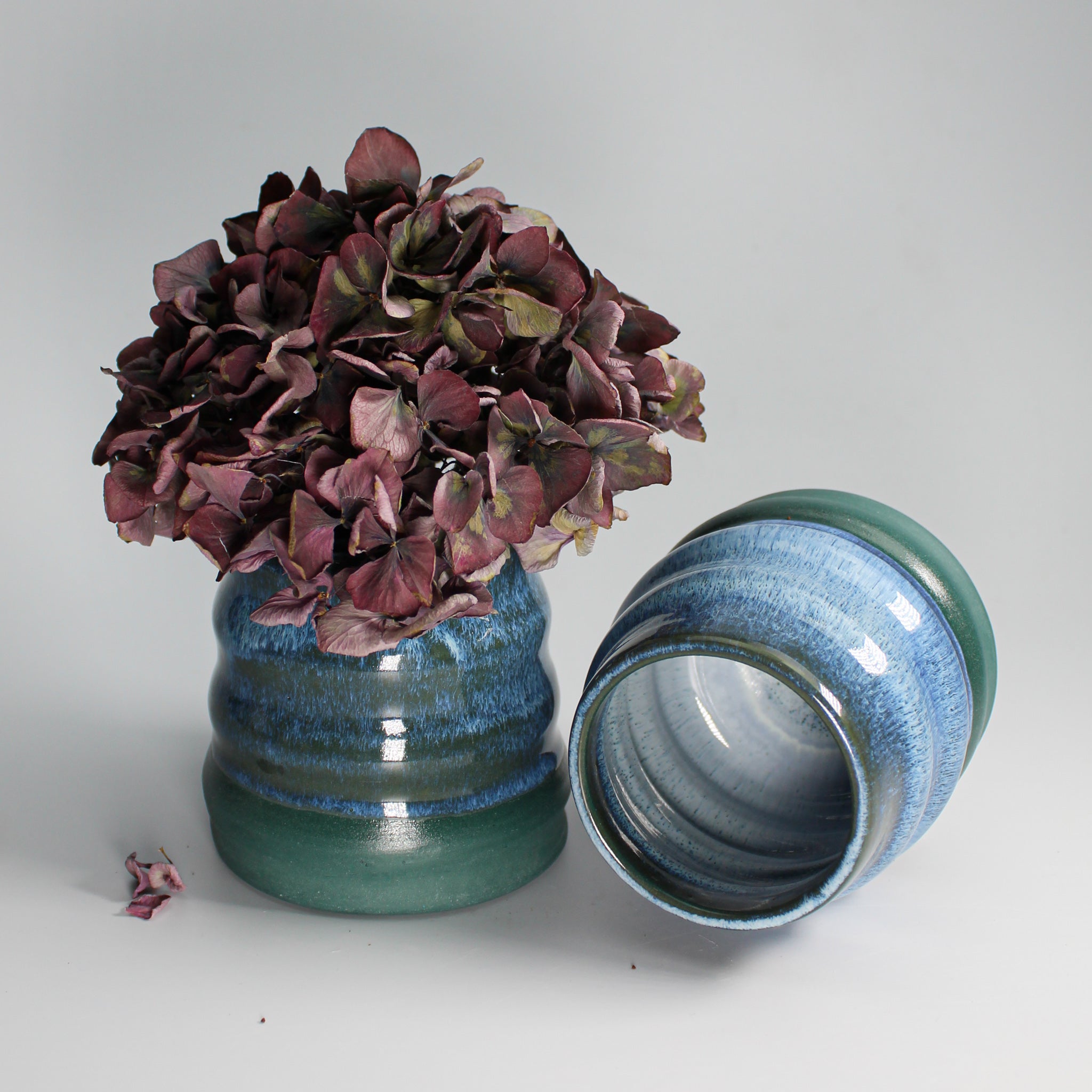 Pair of blue and green mini wobble vases in pottery. One has dried flowers in and the other is lying on its side. 